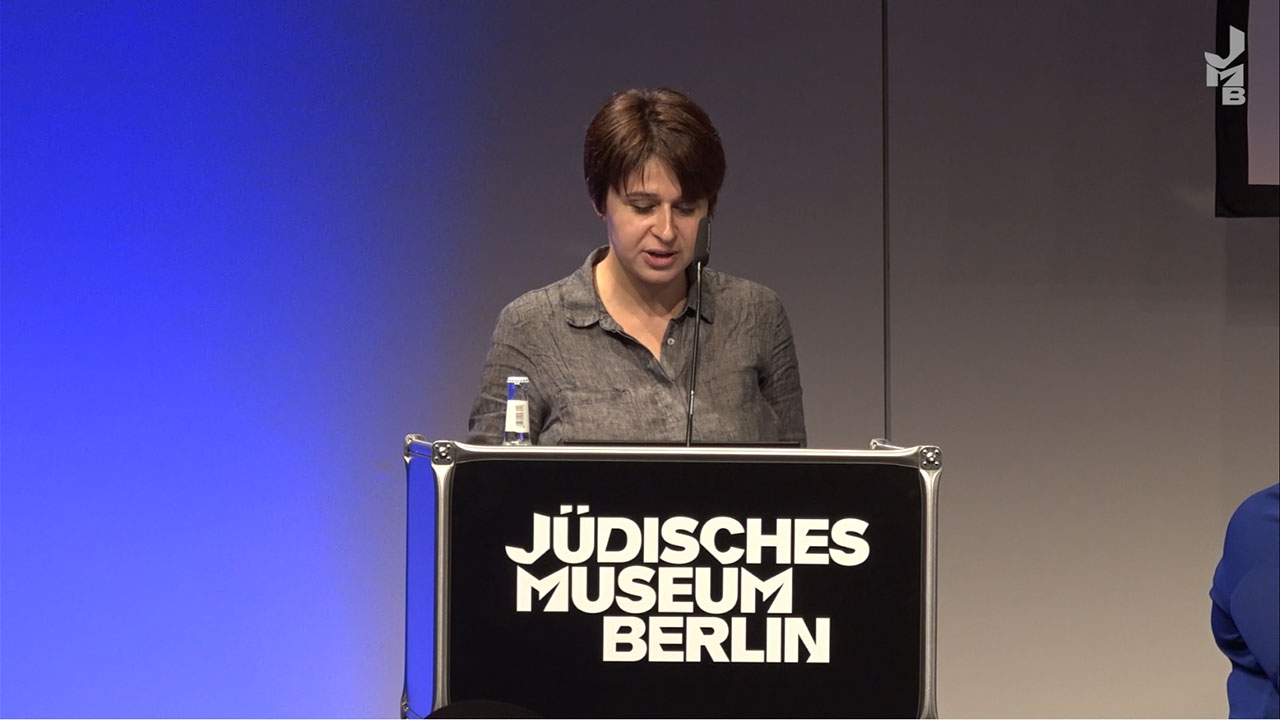 Woman with dark hair stands in front of blue background at black lectern, on which is written Jewish Museum Berlin.