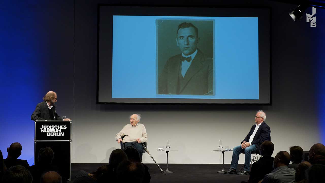 Stage on which two men are sitting, another is at the lectern.