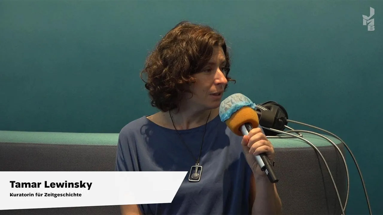 Videostill: Eine Frau mit braunen Locken spricht in ein Mikro, darunter eine Bauchbinde: Tamar Lewinsky, Kuratorin für Zeitgeschichte.
