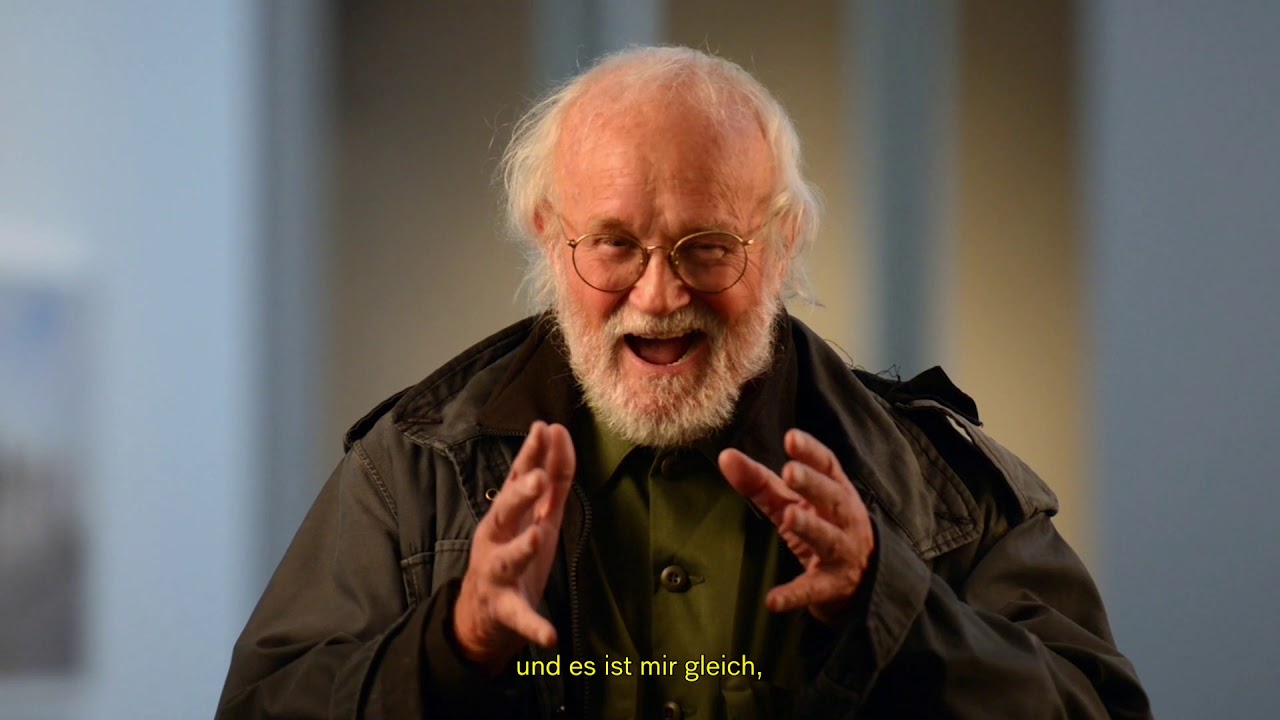 Man with white hair and glasses gestures with both hands.