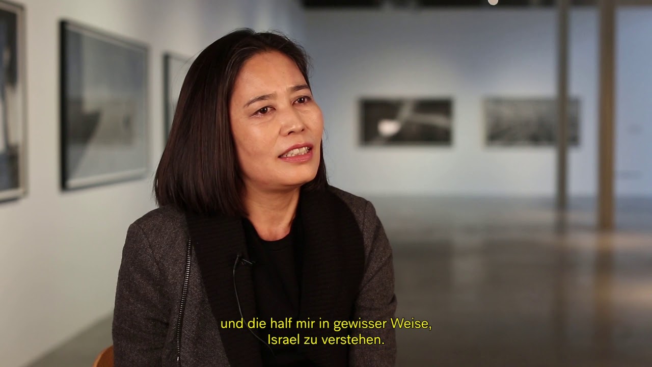 Woman with dark hair sits in an exhibition and gives an interview.
