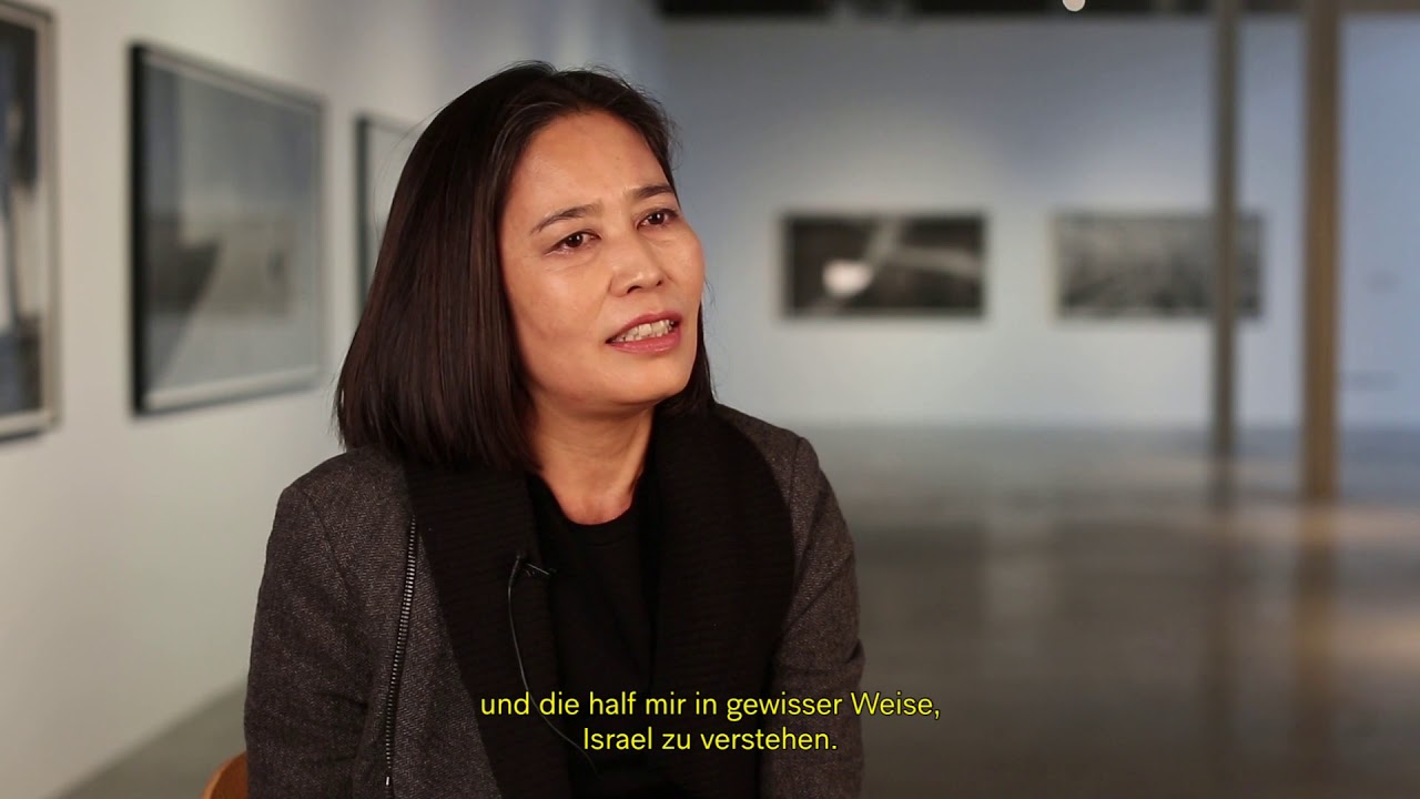 Frau mit dunklen Haaren sitzt in einer Ausstellung und gibt ein Interview.