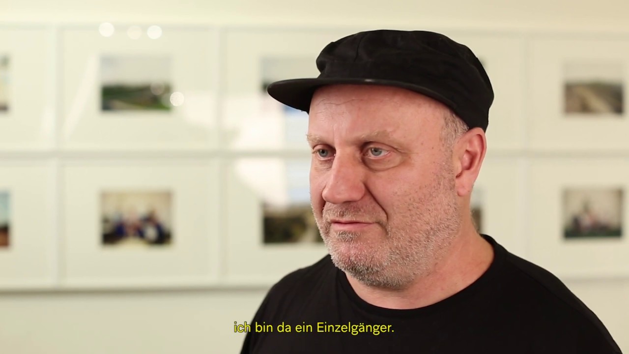 Man with black cap stands in front of pictures in an exhibition.