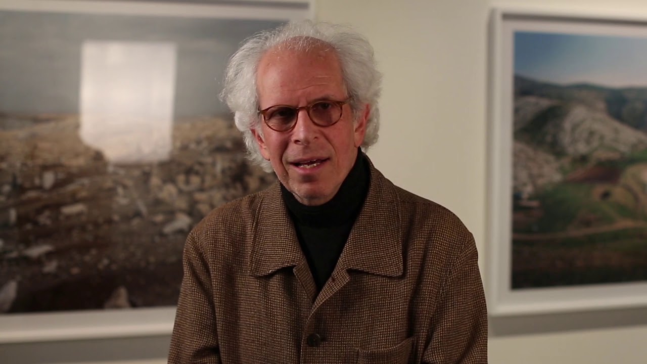 Man with white hair and black glasses speaks into the camera.