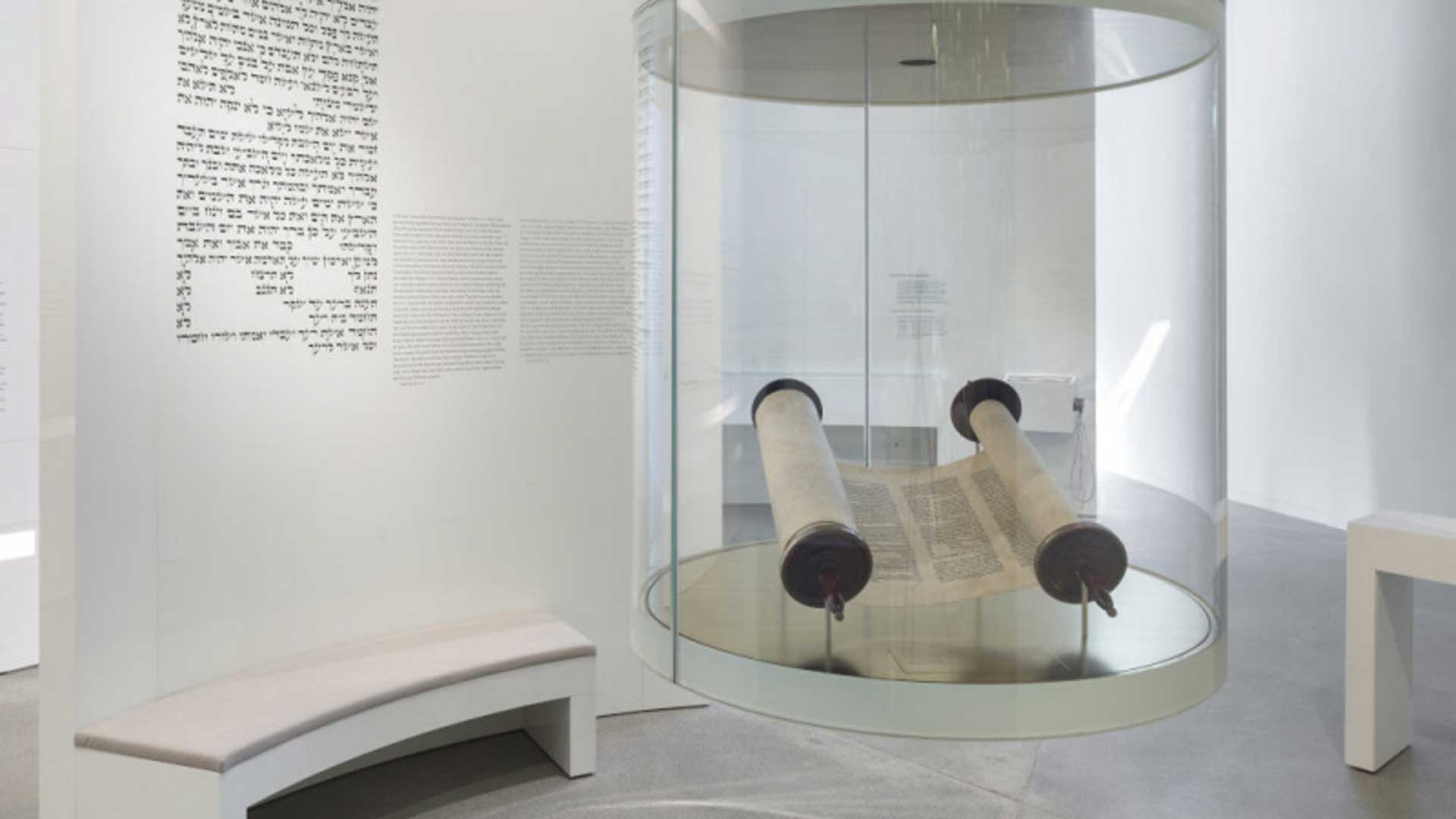 Eine Tora-Rolle ist in einer Museumsvitrine ausgestellt, auf der weißen Wand im Hintergrund steht ein hebräischer Text.