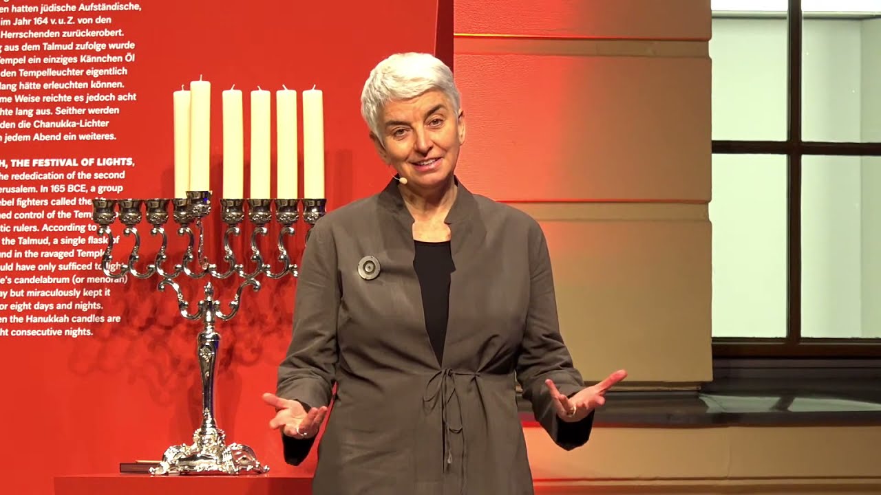 A woman with short gray hair (Hetty Berg) stands in front of a Hanukkah menorah..