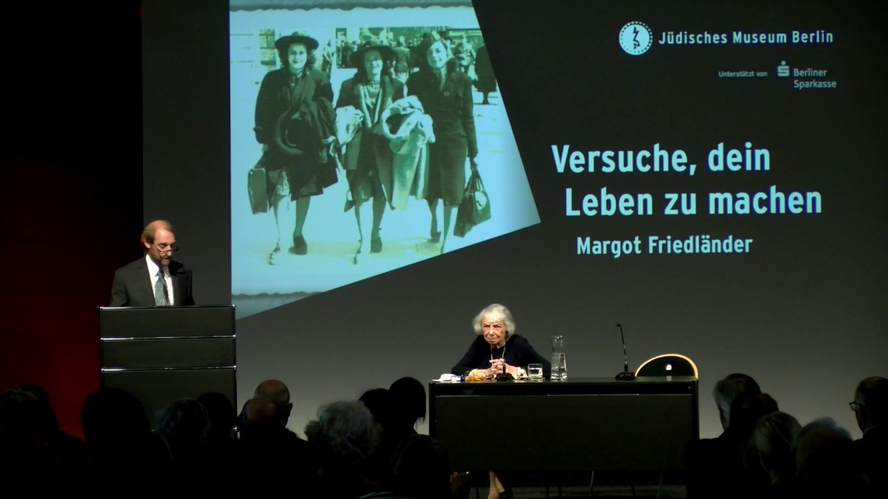 On a podium, Aubrey Pomerance is standing at a lectern (on the left side of the picture), Margot Friedländer is sitting at a table (on the right side of the picture)