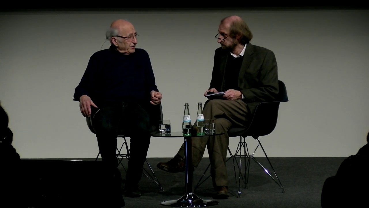 Two men sit talking at a low coffee table: Peter Neuhof on the left, Aubrey Pomerance on the right.