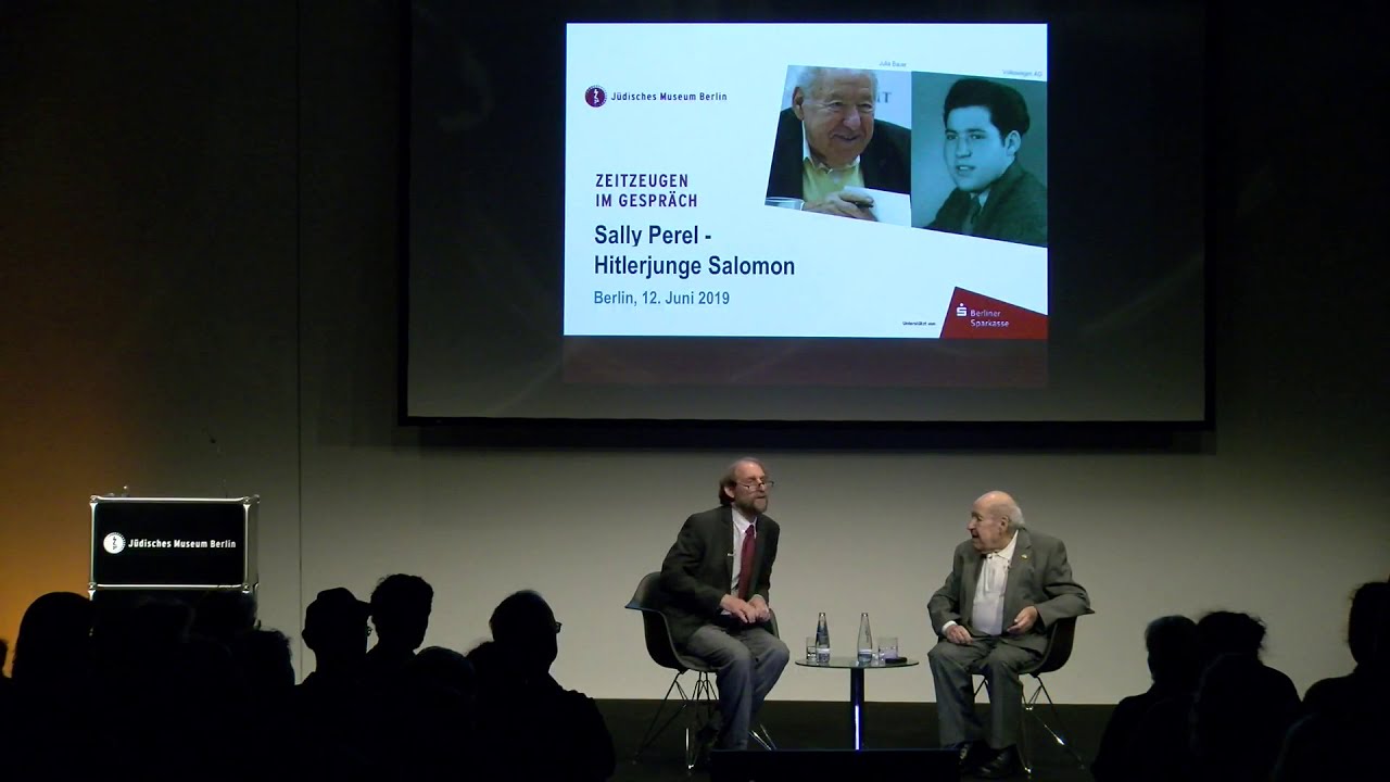 Two men sit talking at a low coffee table: Sally Perel on the right, Aubrey Pomerance on the left.