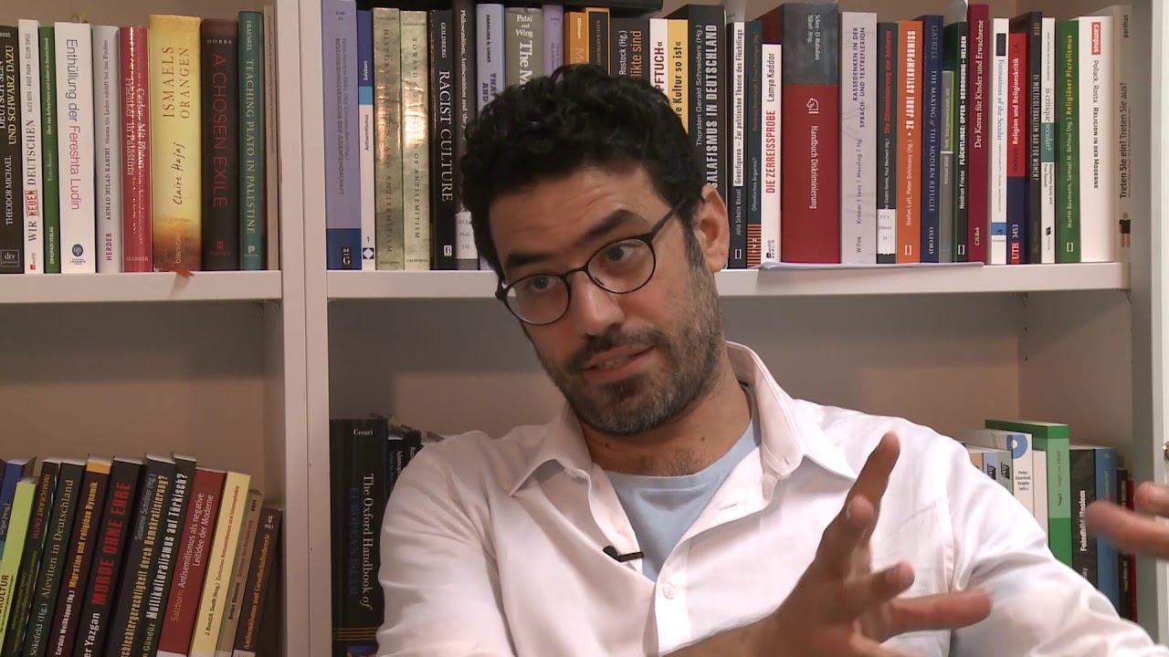 A man in a white shirt is sitting in front of a bookshelf giving an interview.