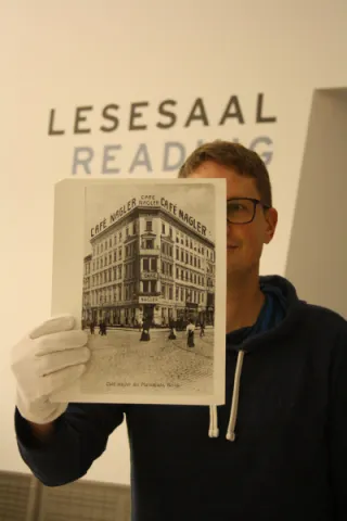 Ein Mann hält eine Schwarz-Weiß Fotografie vor sich. Auf dem Foto ist ein Gebäude abgebildet. 