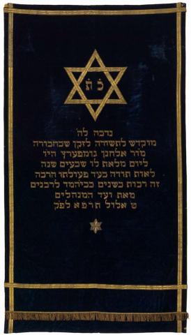Tora-Vorhang aus dunkelblauen Seidensamt mit schlichten Goldstickereien, im Zentrum ein Davidstern, in dessen Mittelfeld die hebräischen Buchstaben „K“ (=Kaw) und „T“ (=Taw) als Abkürzung für „Keter Tora“ (= Krone der Tora) eingestickt sind.