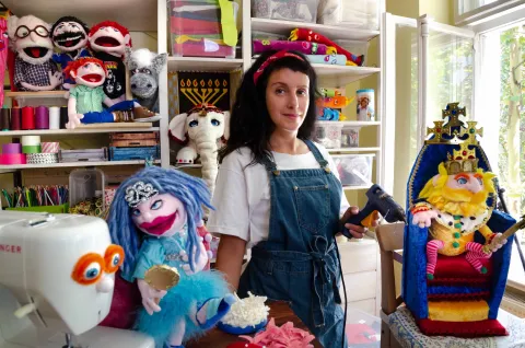 The picture shows a woman wearing dungarees and a white T-shirt. She is surrounded by dolls and a sewing machine.