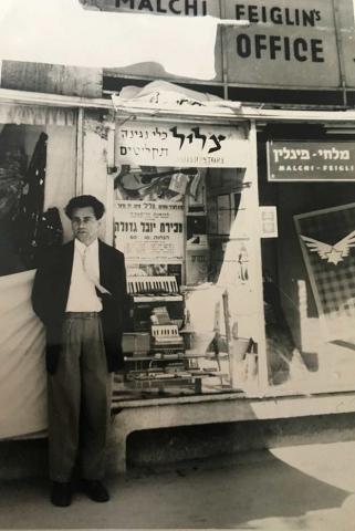Black-and-white photograph of a man in front of a shop window display with musical instruments and Hebrew writing