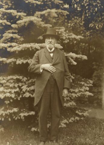 Photo of a man with suit and hat in front of a coniferous tree.