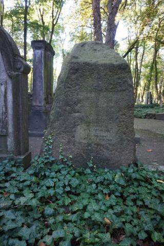 Farbfoto: Grabstein mit stark verwitterter Inschrift, Efeu berankt, im Hintergrund weitere Gräber