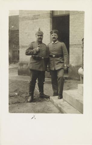 Black-and-white photograph: two soldiers in uniform, one in a spiked helmet and the other in a military cap, outside a building