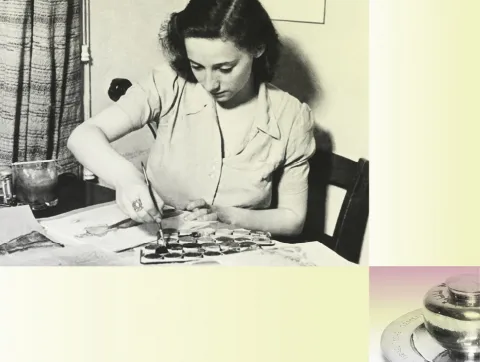 A woman is sitting at her desk covered with colours and brushes. She is painting.