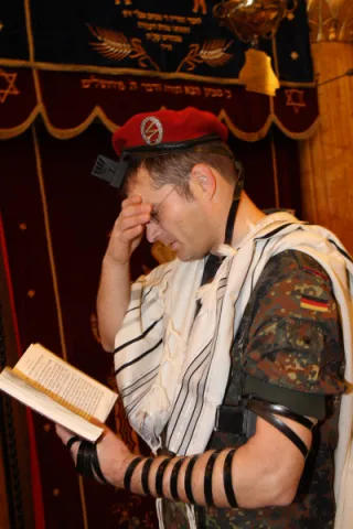 Ein Mann in Bundeswehruniform h&auml;lt in der linken Hand ein aufgeschlagenes Buch, die rechte Hand h&auml;lt er vor seine Augen, sein Blick ist gesenkt auf das Buch. An seinem linken Arm tr&auml;gt er einen j&uuml;dischen Gebetsriemen.