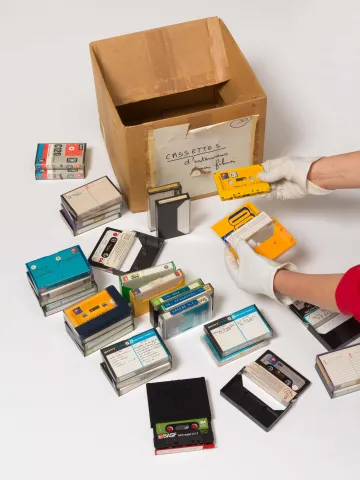 Kiste, Kassetten und zwei Hände. Eine Hand hält eine der Kassetten. 