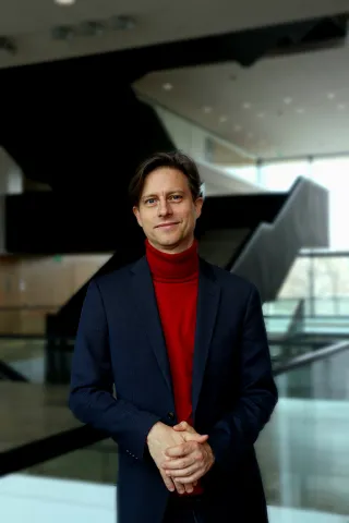 A man in a jacket and red turtleneck sweater stands in a hall and smiles in a friendly manner.