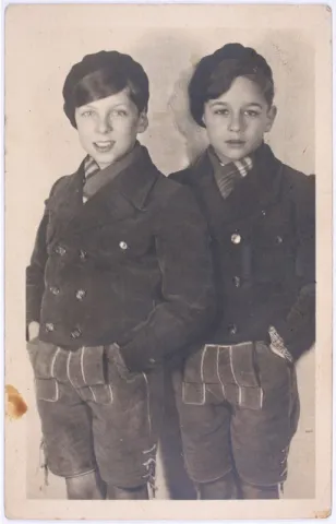 Black-and-white photo. Two boys wearing jackets and shorts stand next to each other and look at the camera.