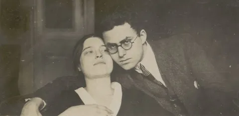 Die historische Fotografie zeigt eine junge Frau, zu deren rechter Seite ein Mann an ihren Kopf gelehnt ist. Er blickt in die Kamera und umfasst die Schultern der Frau mit seinem rechten Arm.