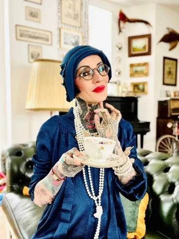 Eine Frau steht in einem Wohnzimmer mit Ledersofa, Klavier und Bildern an den Wänden. Sie trägt ein blaues Kleid, eine lange Perlenkette, Hut und roten Lippenstift.