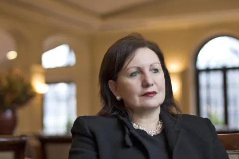 Eine Frau schaut nach rechts. Sie tr&auml;gt dunklen Lippenstift, hat dunkle Haare und tr&auml;gt schwarze Kleidung und Perlenohrringe.