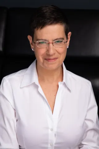 A woman with glasses and a white blouse looks kindly at the camera.
