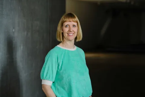 A woman with shoulder-length, blonde hair is smiling and looking directly towards the camera. Her upper body is visible. She is wearinga turquois coloured sweater.