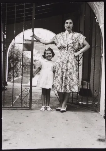 Schwarz-Weiß Bild. Eine Frau mittleren Alter und ein kleines Mädchen stehen neben einem Gittertor vor einem Hauseingang