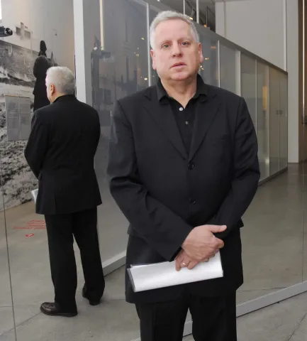 Stehender Mann in schwarzem Anzug vor einer verspiegelten Glasfläche, seine Körperrückseite ist als Spiegelbild zu sehen
