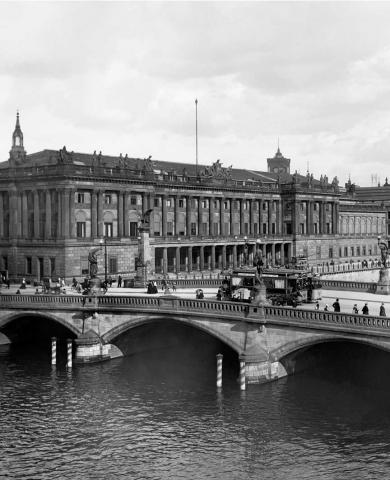 A black and white photograph of a large neoclassical building on a riverbank and a bridge crossing the water. 