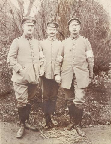 Black and white photograph with three soldiers in uniform frontal and standing in front of a green area.
