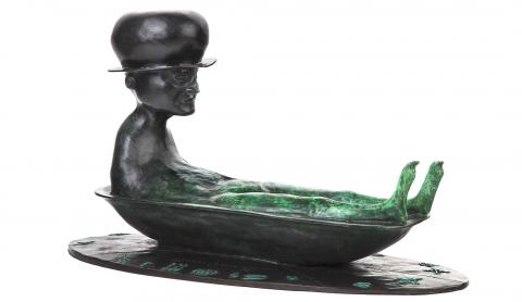 Greenish-black figure made of bronze: a person with a hat in a small bathtub.
