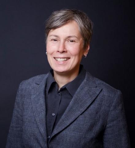 Portrait of a woman with short gray hair and gray blazer in front of a black background. 
