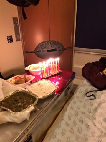 In a sleeping car on the train, a metal suitcase covered by a meal on plastic and aluminum plates, next to it a candelabrum with eight lit candles.