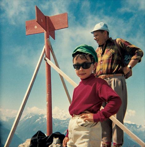 A little boy with sunglasses stands in front of a summit cross, in the background is an older man.