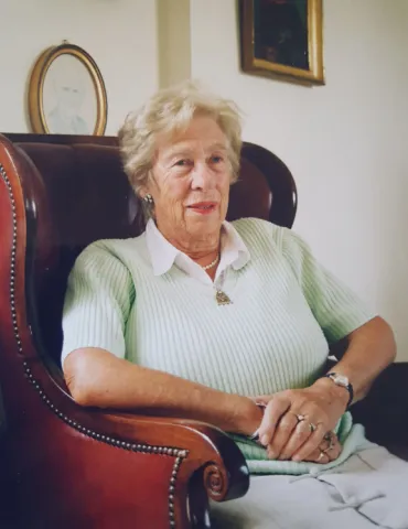 An elderly woman sits in a wing chair, framed pictures on the wall behind her