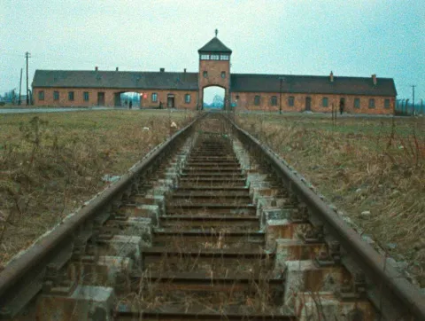 Bahngleise, die auf ein breitgestrecktes, flaches Backsteingeb&auml;ude zuf&uuml;hren. In der Geb&auml;udemitte befindet sich ein Wachturm mit einem Tor f&uuml;r die Durchfahrt ankommender Z&uuml;ge. Es handelt sich um das Lagertor des Vernichtungslagers Auschwitz-Birkenau.