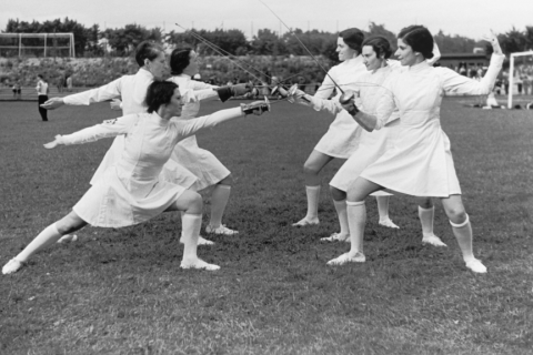 Six women facing each other in white clothes. They pose in a fencing attack position.