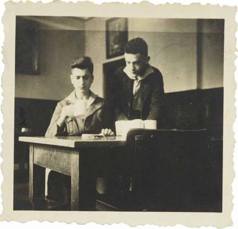 Zwei Jungen an einem Tisch, einer sitzend mit einer Tasse, einer schräg hinter ihm stehend (Schwarz-Weiß-Fo­to)