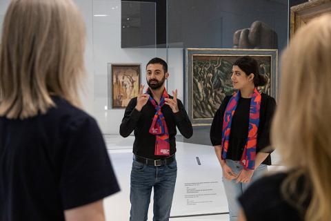 A man stands next to a woman in an exhibition room with paintings and speaks in sign language.