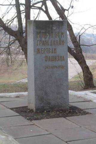 Schlichter grauer, aufrecht stehender Stein mit goldener Inschrift in kyrillischen Buchstaben, im Hintergrund zwei Bäume.