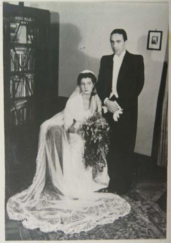 Black-and-white photo of a bridal couple. The bride is seated with the groom standing behind her. A cabinet is visible to the left in what appears to be a living room. 