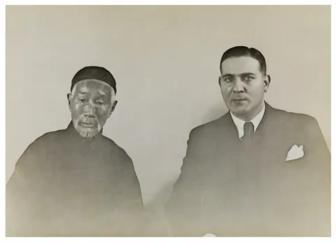 Schwarz-Weiß Bild. Links ein älterer Mann in dunkler Kleidung. Rechts ein mittelalter Mann in einem Anzug. Beide schauen in die Kamera. 