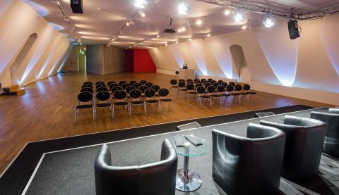 In the empty conference room, large black leather chairs with small round glass tables face rows of empty chairs