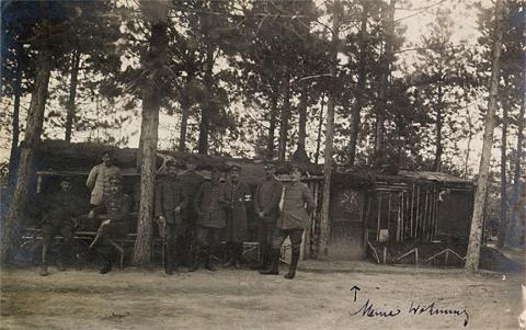 Schwarz-weiß Fotografie mit mehreren uniformierten Soldaten zum Teil sitzend und stehend vor einer Hütte im Wald.