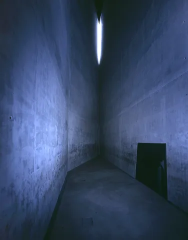 Innenansicht des Holocaust-Turms: Ein enger Raum mit hohen, grauen Betonw&auml;nden. Von oben f&auml;llt etwas Licht ein.